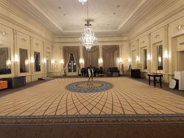 Elegante salão de baile com teto ornamentado, lustre de cristal, tapete bege e dourado, arandelas, espelhos e cortinas elegantes. Duas cadeiras pretas vazias no centro..