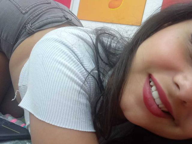 LeilaEvanns est penchée en avant, vêtue d'un t-shirt blanc côtelé ajusté et de shorts gris, avec son sourire aux lèvres rouges et ses cheveux noirs en premier plan, montrant ses fesses rondes..
