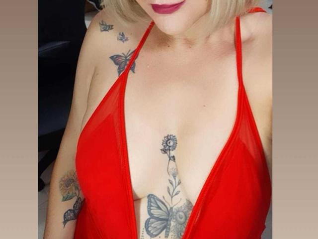 Cexy, con cabello rubio, vestido rojo y tatuajes en el pecho y los brazos, mostrando sus grandes senos y un tatuaje de mariposa debajo de sus pechos. También lleva lápiz labial rojo..