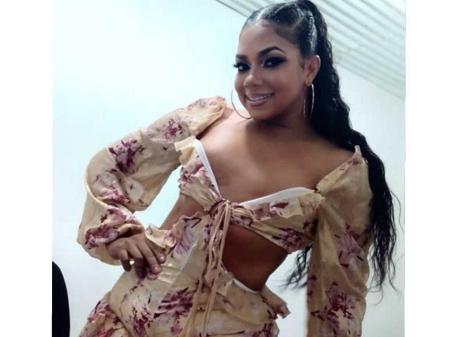 AbellaPhill with long black hair in a ponytail, wearing a floral, off-shoulder, tied-up crop top and matching skirt, smiling, showing her small tits and flat ass.