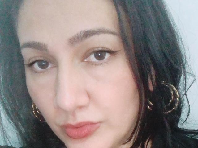 Le visage de QueenVentura, avec sa peau claire, ses yeux marron foncé et ses longs cils, ses lèvres rosées légèrement entrouvertes, et ses cheveux noirs ondulés ornés de boucles d'oreilles dorées, la regardant droit dans l'objectif..
