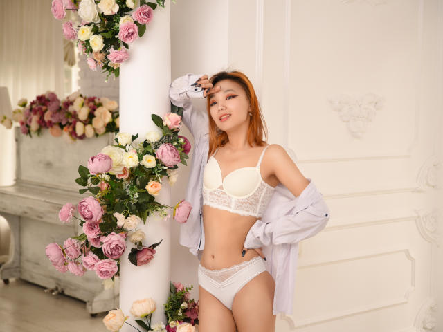 MillaWaves with light brown hair, wearing white lace bra and panties, a loose shirt hanging off her shoulders, standing by a floral-covered column, showing her tits and pussy.