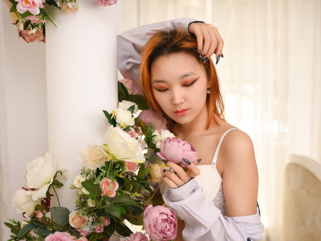 Milla, con i capelli arancioni, che tiene una rosa rosa, indossa un reggiseno bianco e una maglietta grigia, con le braccia alzate sopra la testa, circondata da fiori, luce soffusa, espressione delicata, trucco leggero, molto carina..