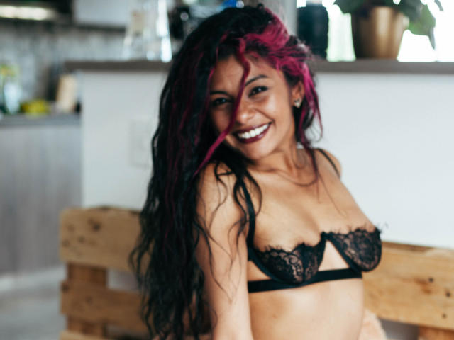 Anna, con capelli rosa e neri, sorridente, indossa un reggiseno di pizzo nero, mostrando piccoli seni, seduta su una panca di legno, con uno sfondo di cucina sfocato, luce naturale, casual, sexy, sicura di sé..