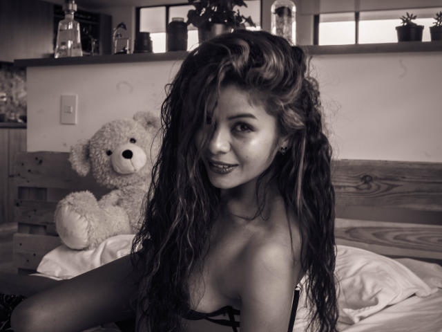 Anna, con i capelli mossi e spettinati, sorride alla fotocamera, indossa un top nero con spalline, che mette in mostra il seno, ed è seduta su un letto con un orsacchiotto di peluche accanto a lei..