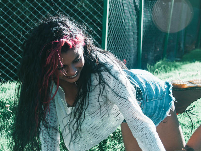 AnnaMesht a quattro zampe, sorridente, con capelli ricci scuri e riflessi rosa, indossa una maglia bianca di maglia e pantaloncini di jeans blu, luce solare che filtra attraverso una recinzione in rete metallica, terreno erboso..