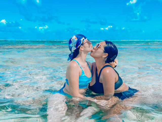 PaolaYKatty with blue hair in a headscarf, wearing a blue bikini, kisses another PaolaYKatty with black bikini, in clear shallow water, both with wet hair and sunglasses.