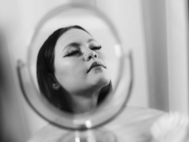 Il volto di Jazmin Wells riflesso in uno specchio, occhi chiusi, trucco leggero, capelli scuri, labbra leggermente dischiuse, luce soffusa, bianco e nero, specchio in primo piano, riflesso leggermente sfocato..