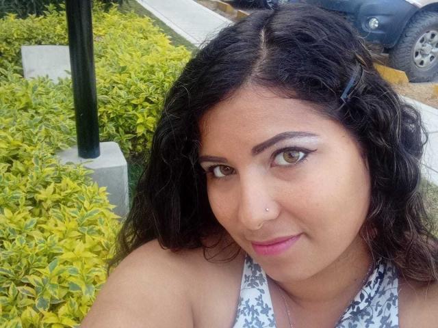 ArtemisaRoses tem cabelos pretos cacheados, um pequeno piercing no nariz, olhos castanhos e batom rosa, vestindo uma blusa branca com alças e um padrão preto, e está em pé perto de arbustos verdes..