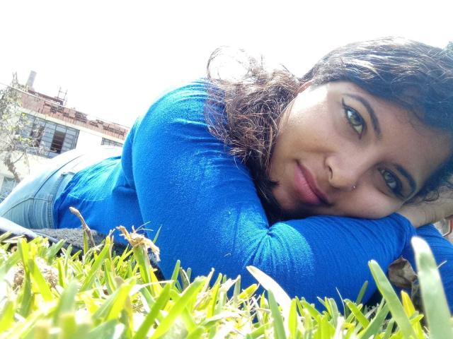 ArtemisaRoses sdraiata sull'erba, con il viso tra le mani, indossa una camicia a maniche lunghe blu, ha gli occhi verdi, un piccolo piercing al naso, capelli castani ricci, è una giornata di sole, ha un sorriso dolce, il seno è visibile, appare rilassata..