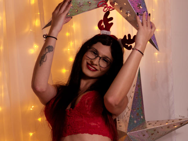 Maddymayer with long black hair, red lace bra, glasses, and antler headband, smiling, holding star-shaped decorations, arms raised, tattoo on left arm, yellow fairy lights.