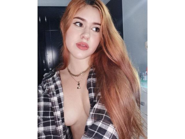 Alessa Clark, con lunghi capelli arancioni, indossa una camicia a quadri nera e bianca aperta, che lascia intravedere i suoi piccoli seni nudi, e guarda la fotocamera con le labbra tumefatte, indossando una collana d'oro..