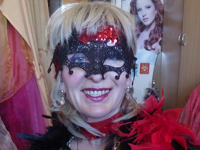 ElizaWendy, avec ses cheveux blonds, un masque en dentelle noire, des paillettes rouges, une plume rouge, souriante, portant des plumes rouges et noires, du rouge à lèvres rose, des yeux bleus, et une écharpe rouge sur une photo..