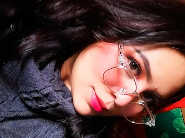 Samantha Velez, con cabello oscuro y ondulado, usando gafas transparentes, lápiz labial rosa y una camisa gris oscuro con textura, acostada, sonriendo suavemente a la cámara..