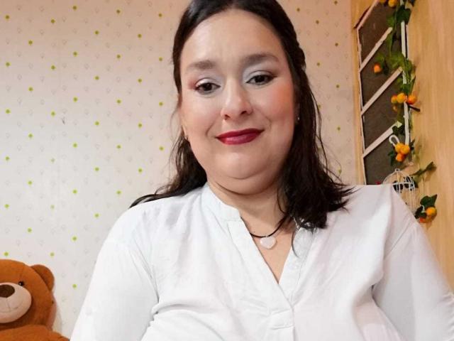 LadySelene has chubby tits, wearing a white shirt, red lipstick, and a necklace, smiling with dark hair, in a room with a teddy bear and orange decor.