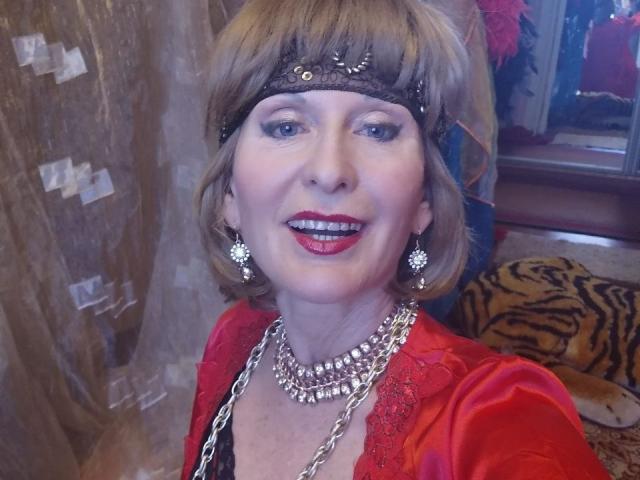 ElizaWendy, avec ses courts cheveux bruns et son rouge à lèvres rouge, portant un bandeau noir, une robe en dentelle rouge et un collier en argent, sourit à l'appareil photo. Ses seins sont petits, et elle est séduisante..