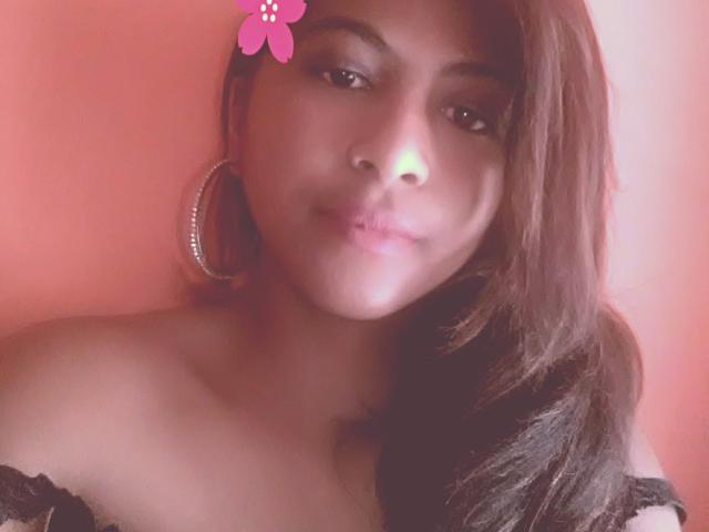 Prianka with brown hair, wearing large hoop earrings and a black off-shoulder top, has a cute pink flower sticker on her forehead, pouting slightly.