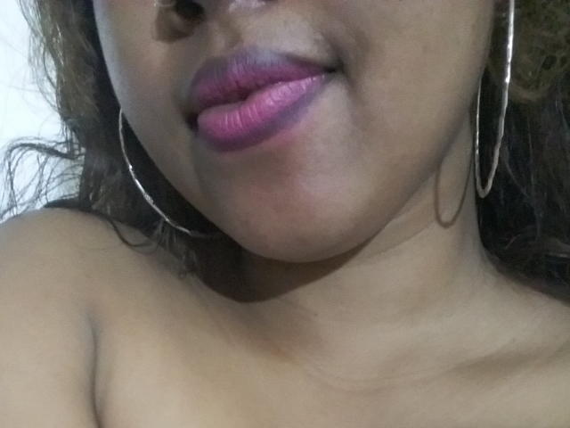 Prianka's face, focusing on her pink glossy lips and dark brown skin. She's wearing large silver hoop earrings and her brown curly hair is partially visible.
