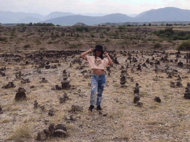AlisonWester with dark hair, wearing a tan crop top and ripped blue jeans, stands in a rocky desert, hands in hair, mountains in the background, and small rock formations.