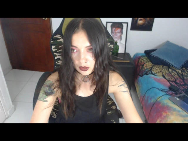 PollyAnnaX tiene el cabello largo y castaño, lápiz labial oscuro y tatuajes en los brazos. Lleva una camiseta negra y una silla con estampado de camuflaje, además de un piercing en la nariz y un collar de aros..