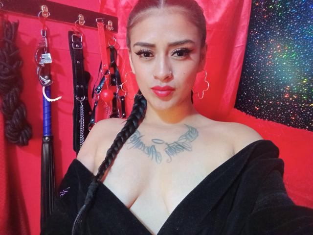 Sexy en sensueel, met gevlochten haar, rode lippenstift, oorringen, een zwarte top met open schouders, diepgang in de hals en een tatoeage op de borst, staand in een kamer met BDSM-attributen..