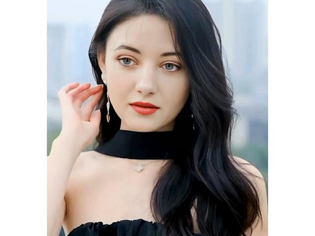 SweetttAngel has pale skin, dark wavy hair, red lipstick, black choker, and is touching her ear with her right hand. She's wearing a strapless black top.