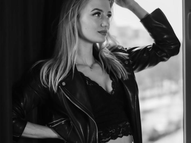 Black-and-white photo of LenaaDoll with long blonde hair, wearing a leather jacket and lace top, hand in hair, looking out a window, showing her tits and subtle ass.