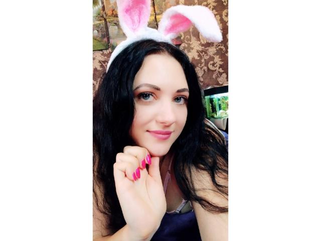 SweetttAngel con una mano con le unghie rosa, capelli neri ondulati, rossetto rosa, orecchie da coniglio bianche, sorridente, leggero blush rosa, pelle chiara, carina, sexy, indossa una maglietta bianca, tiene in mano un telefono..