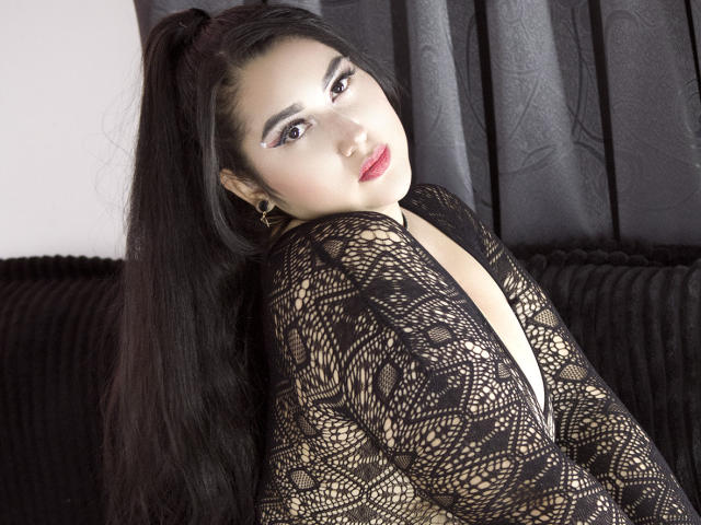 Larysa Tayloor, com cabelos pretos longos presos em um rabo de cavalo, vestindo uma blusa preta de renda com um decote em V profundo, olhando para trás com batom vermelho e brincos de ouro..