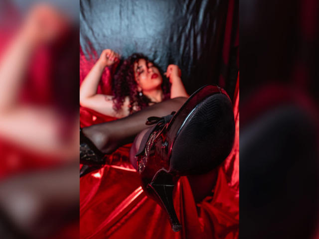 ScarlettBigAss ligt op rode satijn, met krullend haar, gekleed in zwart kant en doorschijnende kousen, met de benen omhoog, zwarte hoge hakken zichtbaar, armen gespierd, verleidelijk kijkend, in een donkere kamer..