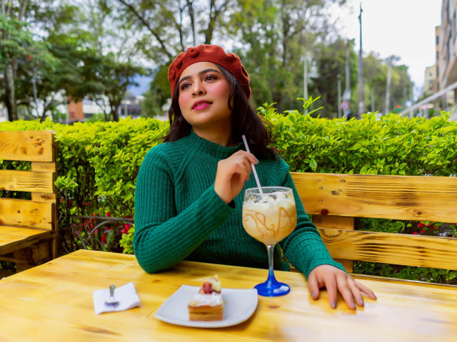 SamanthaVelez with brown hair, green sweater, and red headband, sipping iced coffee with a straw, eating cake, and resting hand on wooden table, looking confident.