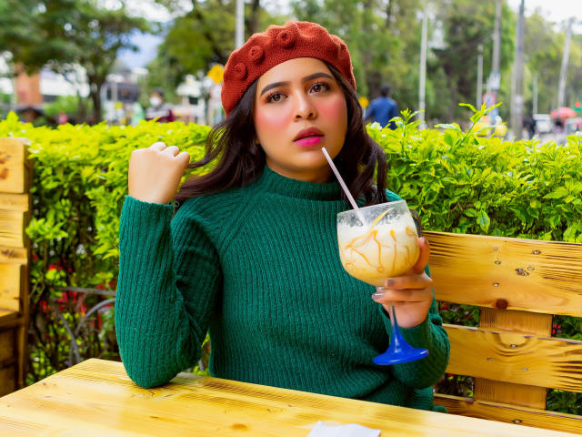 Samantha Velez, con cabello oscuro, boina roja, suéter verde, sosteniendo una bebida con pajita, sentada en una mesa de madera, mirando a la cámara, en un entorno exterior soleado, con textura de madera, y arbustos verdes de fondo..