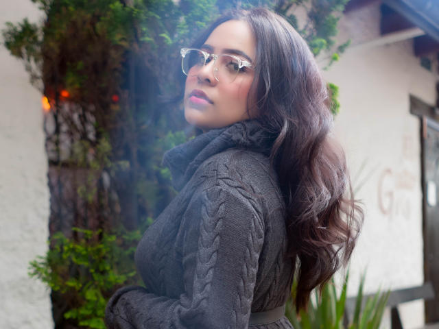 Samantha Velez, con cabello largo, oscuro y ondulado, usando gafas transparentes y un suéter de cuello alto gris oscuro de punto grueso, mirando por encima del hombro, mostrando ligeramente su trasero..