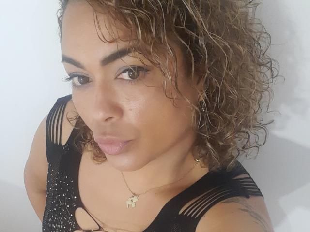 Solmary, con el pelo castaño rizado, vestida con una blusa negra con detalles recortados, labios carnosos, ojos marrones y un pequeño collar dorado con un colgante, mirando hacia la cámara..
