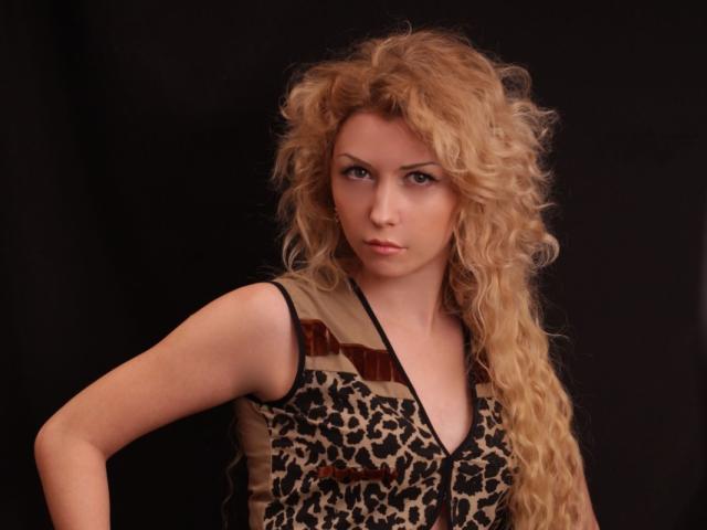 Boginya hat langes, lockiges blondes Haar und einen ernsten Gesichtsausdruck. Sie trägt ein Oberteil mit Leopardenmuster in braunen und schwarzen Streifen auf schwarzem Hintergrund..