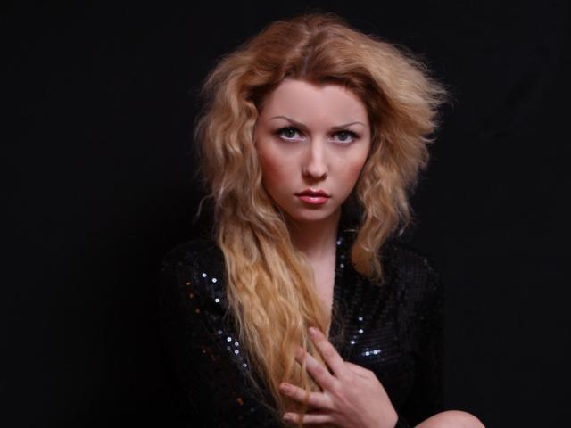 Una donna, Boginya, con lunghi capelli biondi ondulati, indossa una top nera con paillettes, ha un'espressione seria, una mano che tocca il petto, su uno sfondo scuro. Seno, capezzoli, glutei non visibili..