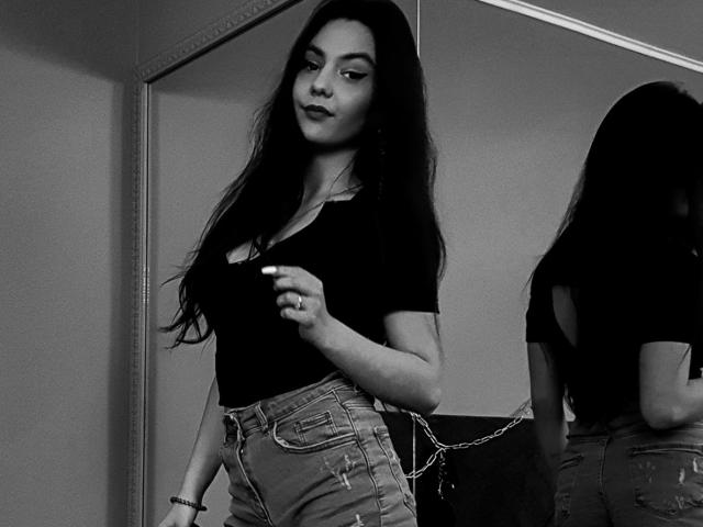Foto em preto e branco de AshlyChloe com cabelos longos e escuros, vestindo uma blusa preta justa e jeans de cintura alta, em frente a um espelho, mostrando seu traseiro no reflexo..
