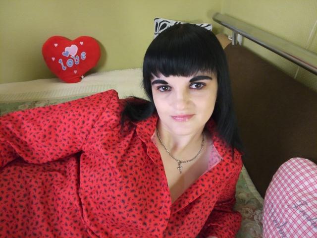 XLylyusX met zwart haar en een pony, die een rood stippen shirt draagt, ligt op een bed, glimlacht, draagt een kruisbeeldketting, is schattig, heeft kleine borsten, bruine ogen, en er is een rode hartdecoratie op de achtergrond..