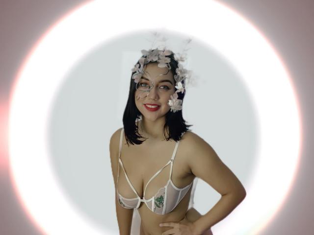 SquirtinGamy with black hair, wearing white floral headpiece, sheer bra with visible tits, smiling, red lipstick, hands on hips, soft white ring light behind her.