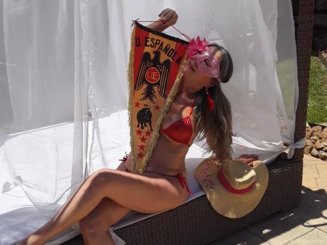 EvaRodinson in red bikini, holding Spanish flag, pink mask, and straw hat, sitting on wicker lounge, white canopy, tan skin, long brown hair, visible ass, sunny.