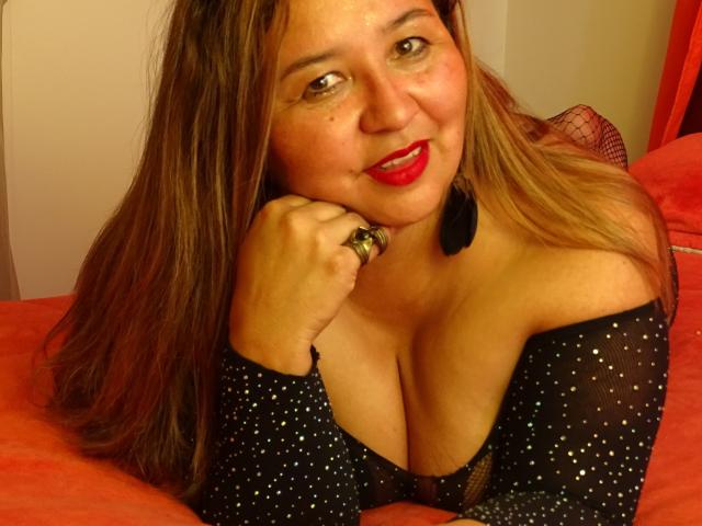 AmbarSimmons with red lipstick, smiling, wearing a sparkly black top, showing her tits, long brown hair, black earrings, ring, and red bed. Her hand rests on her chin.