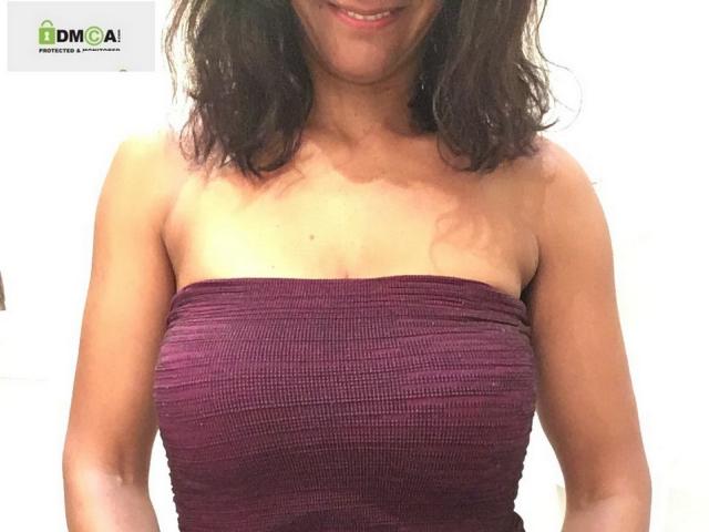 LaetitiaFun with wavy brown hair, smiling, wearing a tight, strapless, purple ribbed dress, showing her small, perky boobs. Her tan skin is visible on her upper chest.