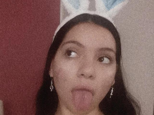ElizabethBlizz with dark hair, wearing a white bunny hat, sticking out her tongue, looking to the side, wearing dangling earrings, has a light brown skin tone, and a cute face.