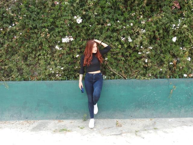 IrisJons com cabelo ruivo, cropped preto, jeans azuis, tênis brancos, segurando um celular, em pé em frente a uma parede verde com flores brancas, com a mão no cabelo, tatuagem visível no braço esquerdo..