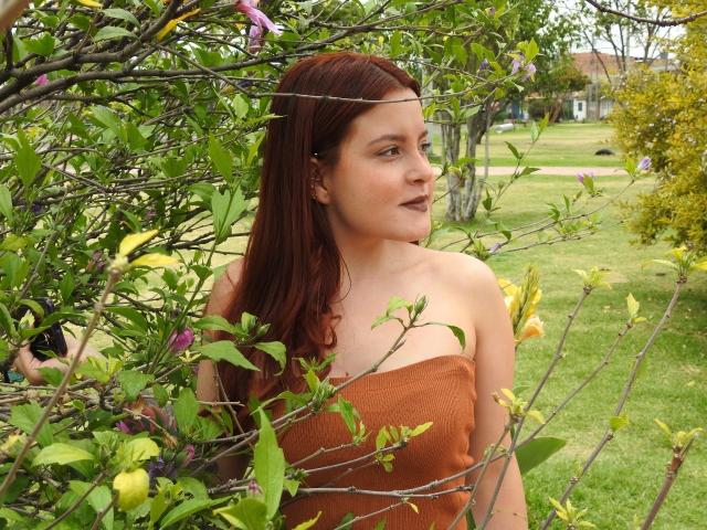 Eine Frau namens Iris Jons mit roten Haaren, mittlerer Brustgröße, in einem braunen Top ohne Träger, steht inmitten von grünen und gelben Blumen, blickt nach rechts, mit leicht geöffnetem Mund und einem ruhigen Gesichtsausdruck..