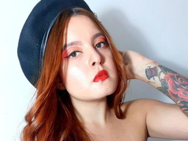IrisJons with red lipstick, orange eyeshadow, and a black beret, showing a tattooed arm, has wavy red hair and a serious face, her tits are slightly visible.
