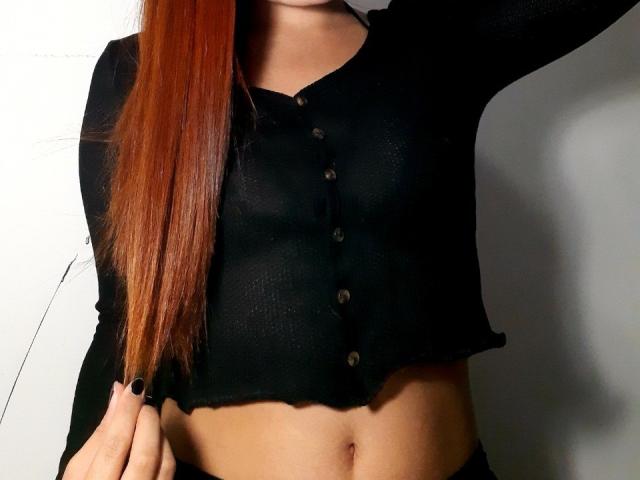 IrisJons with long red hair, black button-up shirt, showing midriff, black nail polish, arm raised, shirt slightly cropped, exposing her bare belly and navel.