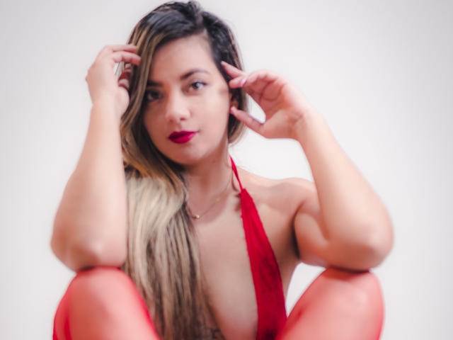 AbrilClark with red lips, blonde and brown hair, wearing a red halter top, sitting with knees up, hands in hair, looking at camera, showing small tits.