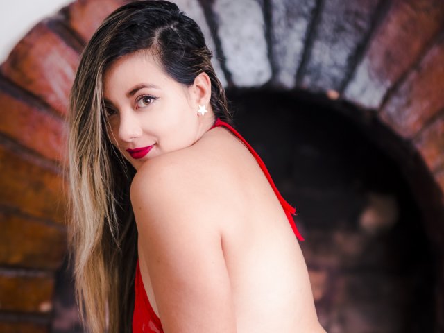 AbrilClark with long blonde hair, red lipstick, and red lingerie, looking over her shoulder, showing her bare back and small white star earrings, smiling. Her tits are partially visible.