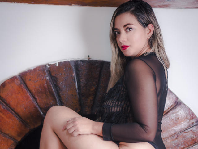 AbrilClark with blonde hair, red lipstick, black sheer dress, sitting on a curved wooden seat, looking back, legs crossed, hands on knees, showing her pussy, brown eyes, confident.